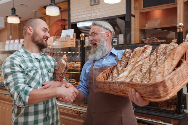 Cheerfu hombre estrecharme la mano con el anciano panadero después de comprar pan fresco en su tienda