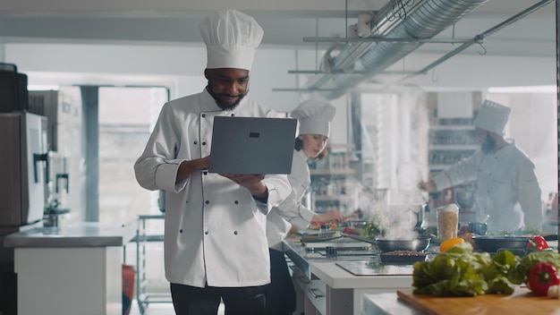 Foto chef afroamericano cocinando receta culinaria con laptop, haciendo preparaciones de comida en la cocina del restaurante. cocinero masculino haciendo plato profesional con comida y utensilios gourmet, servicio de gastronomía.