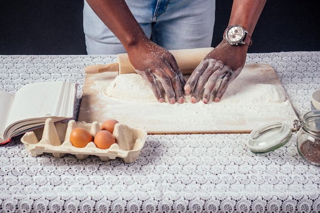 El chef afroamericano se jacta de las manos de repostería en harina para hornear las manos de los hombres masa con rodillo hornea pasteles los huevos y el libro de recetas en la mesa horneado casero