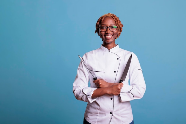 Chef afroamericano con uniforme sonriente mirando a la cámara sosteniendo un cuchillo y un afilador con los brazos cruzados. Cocinera profesional con utensilios de cocina en estudio con fondo azul.