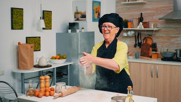 Chef anciano de pie en la cocina en casa y cocinando con productos de panadería de disfrute de harina. Cocinero profesional aplaudiendo y polvo volar espolvorear rew ingredientes sobre la masa preparar brea tradicional