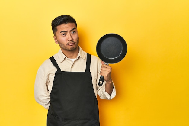 Chef asiático segurando uma panela fundo de estúdio amarelo confuso sente-se duvidoso e inseguro