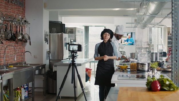 Chef auténtico filmando un programa de cocina en línea en la cocina del restaurante, explicando las preparaciones de recetas culinarias. Cocinera en uniforme grabando video gastronómico para hacer un plato gourmet profesional.