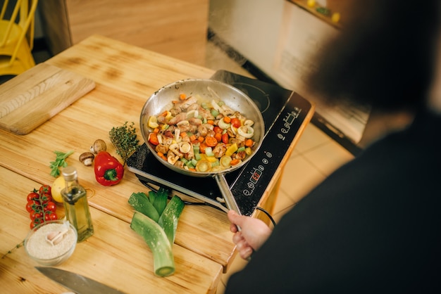 Foto chef barbudo cocinando carne con verduras en la sartén en la cocina.