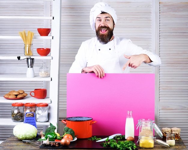 Chef barbudo feliz cocinero señalando con el dedo a la tabla en blanco para anunciar el menú chef en uniforme con vacío