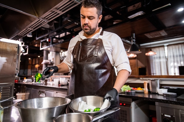 Chef barbudo de mediana edad en delantal y guantes de pie en la estufa y cociendo brócoli de la olla en la cocina del restaurante