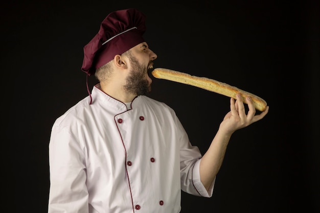 Chef bonito barbudo homem de uniforme morde baguete, cozinheiro padeiro masculino