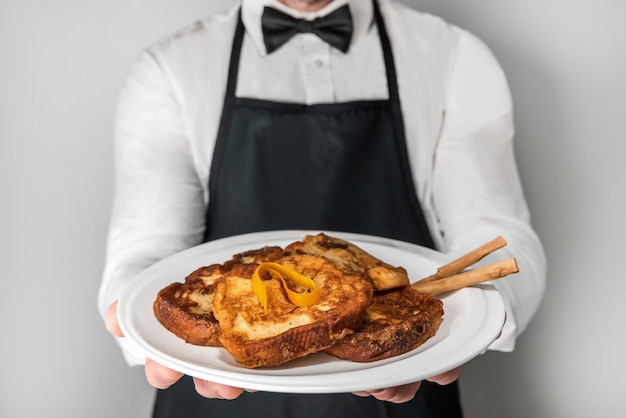 Un chef camarero en delantal y pajarita mostrando un plato de torrijas gourmet español consumido dulce Pascua