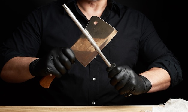 chef en una camisa negra y guantes de látex negro afilar un cuchillo de cocina en un afilador de hierro con un mango por encima de la mesa de baja clave de cerca