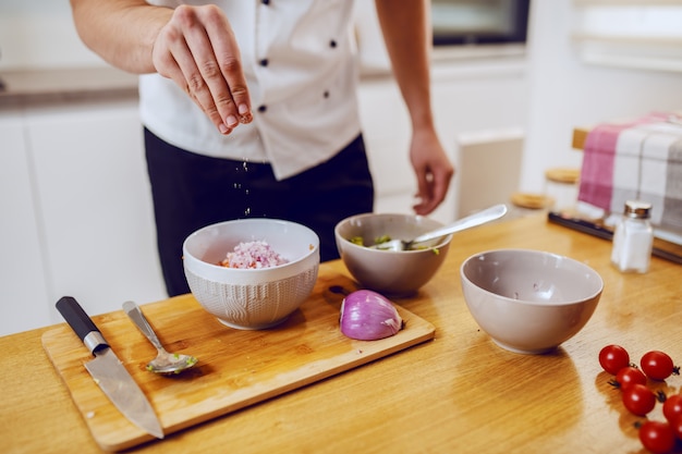 Chef caucásico dedicado en uniforme de pie en la cocina y agregar sal en un tazón con verduras.