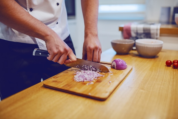 Chef caucásico en uniforme de pie en la cocina doméstica y cortar la cebolla