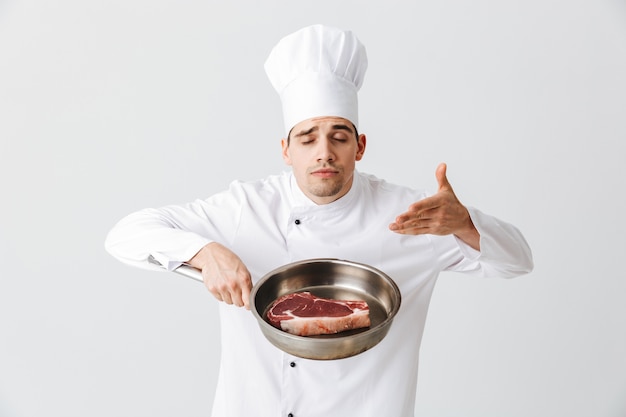 Chef cocinero alegre vistiendo uniforme mostrando una sartén con un filete de ternera cruda aislado sobre la pared blanca