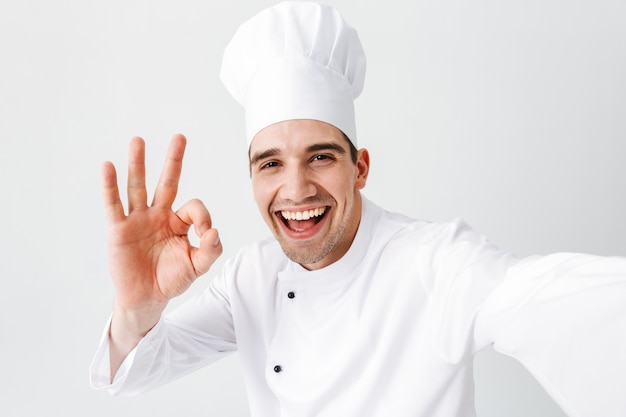 Chef cocinero feliz vistiendo uniforme que se encuentran aisladas sobre una pared blanca, tomando un selfie, mostrando ok