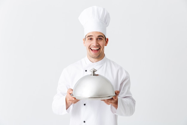 Chef cocinero hombre emocionado vistiendo uniforme con cloche aislado sobre pared blanca