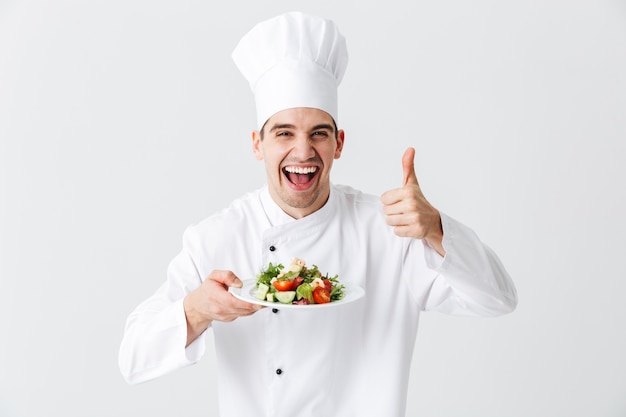 Chef cocinero hombre emocionado vistiendo uniforme mostrando ensalada verde fresca en un plato aislado sobre la pared blanca