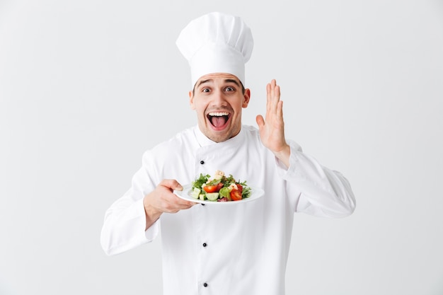Chef cocinero hombre emocionado vistiendo uniforme mostrando ensalada verde fresca en un plato aislado sobre la pared blanca