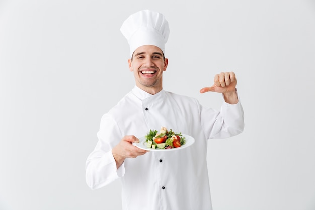 Chef cocinero hombre emocionado vistiendo uniforme mostrando ensalada verde fresca en un plato aislado sobre la pared blanca