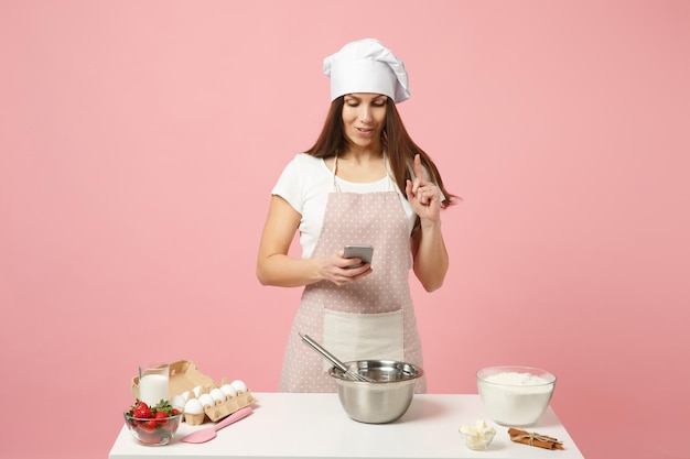 Chef cocinero repostero o panadero con camiseta blanca, sombrero de chef toque pastel de cocina o cupcake en la mesa usando teléfono móvil aislado en un fondo rosa pastel en el estudio. Concepto de comida de espacio de copia simulada