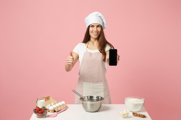 Chef cocinero repostero o panadero con camiseta blanca, sombrero de chef toque pastel de cocina o cupcake en la mesa usando teléfono móvil aislado en un fondo rosa pastel en el estudio. Concepto de comida de espacio de copia simulada