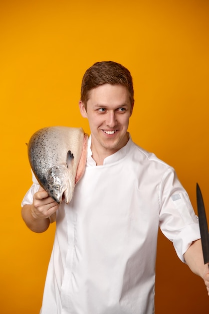 Chef cocinero sosteniendo un gran pez salmón atlántico en su hombro y cuchillo aislado sobre fondo amarillo