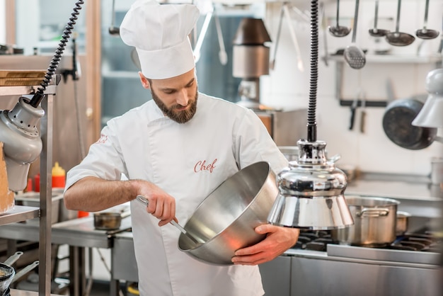 Chef cocinero en uniforme mezclando ensalada en el recipiente de metal en la cocina del restaurante