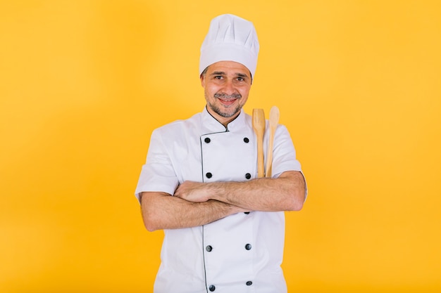 Chef cocinero vistiendo gorro de cocina blanco y chaqueta con cuchara de madera y tenedor y brazos cruzados, sobre fondo amarillo