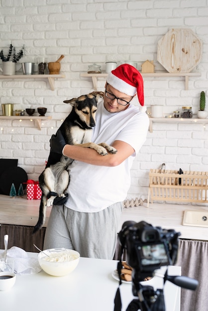 Chef com chapéu de Papai Noel segurando seu cachorro nos braços, cozinhando na cozinha e fazendo vídeo