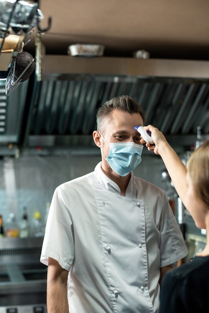 Chef contemporáneo en uniforme y máscara protectora mirando a la mujer con termómetro midiendo la temperatura de su cuerpo antes de trabajar en la cocina