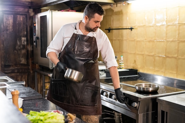 Chef corpulento en delantal de cuero eligiendo la temperatura de la estufa mientras la enciende en la cocina comercial