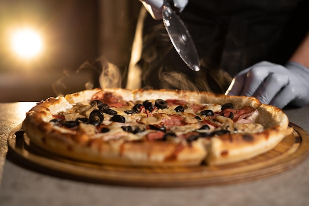 El chef corta pizza caliente con queso y salami con un cuchillo sobre una tabla de madera.