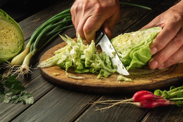 Chef corta repolho fresco com faca para salada em uma mesa de cozinha com legumes frescos