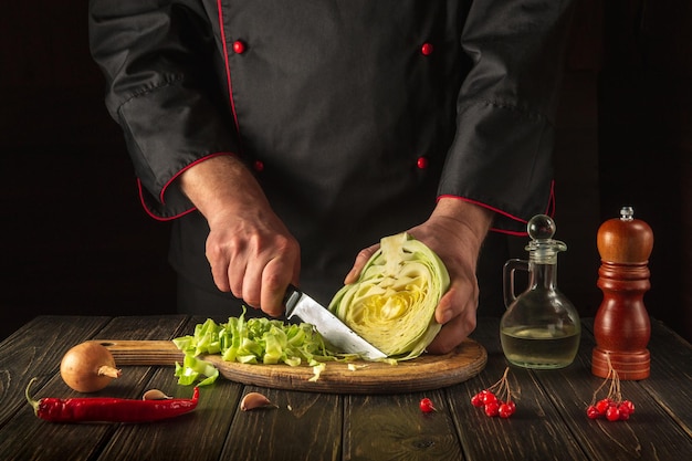 Chef corta repollo fresco con cuchillo para ensalada en una mesa de cocina con verduras frescas