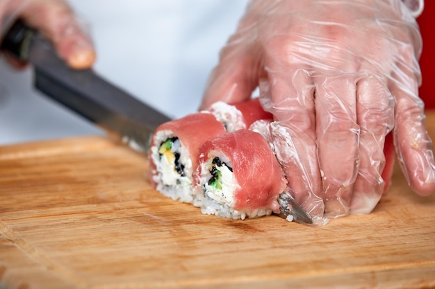 Chef corta rollos de sushi con salmón rojo con un cuchillo en una tabla de madera Chef con guantes transparentes trabaja