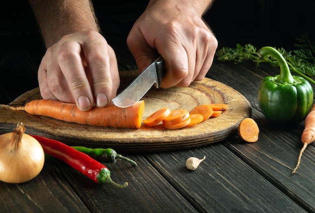 El chef corta la zanahoria en trozos pequeños en una tabla de cortar para preparar un almuerzo o una cena vegetariana Conjunto de verduras en la mesa de la cocina