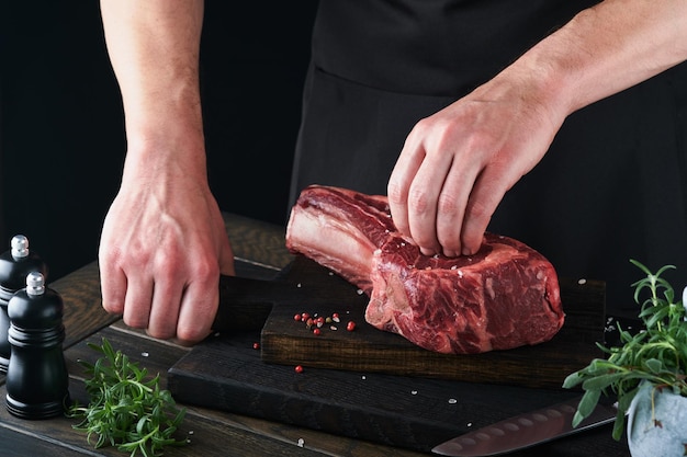 Chef cortando bife As mãos do homem seguram o bife cru Tomahawk na tábua de corte de madeira rústica sobre fundo preto Cozinhando receitas e comendo o conceito Foco seletivo