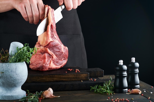 Chef cortando filete de ternera Mans manos sostienen bistec crudo Tomahawk en tabla de cortar de madera rústica sobre fondo negro Recetas de cocina y concepto de alimentación Enfoque selectivo