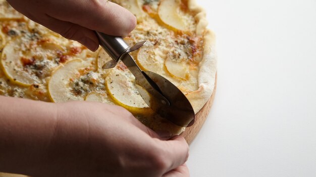 Chef cortando pizza de frutas de pera con un cuchillo sobre una tabla de madera en el primer plano de la mesa blanca