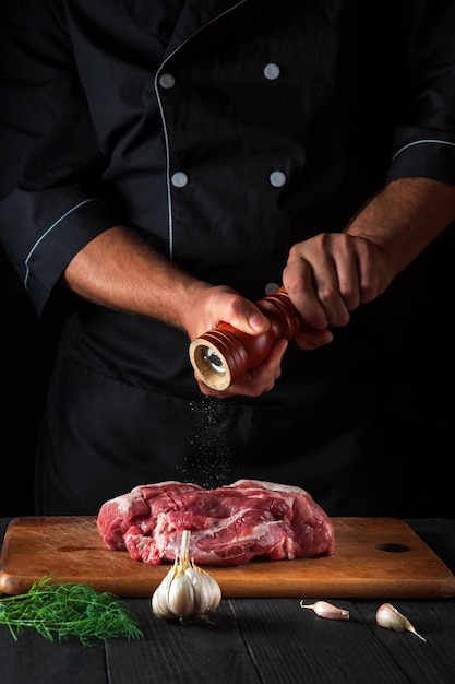 Chef cozinhando filé de carne crua e adicionando pimenta ou pimentão para marinada em fundo preto Ambiente de trabalho na cozinha no restaurante ou café