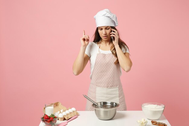 Foto chef cozinheiro confeiteiro ou padeiro em t-shirt branca, chapéu de chefs de toque cozinhar bolo ou cupcake na mesa falando no celular isolado em fundo rosa pastel em estúdio. mock up conceito de comida de espaço de cópia