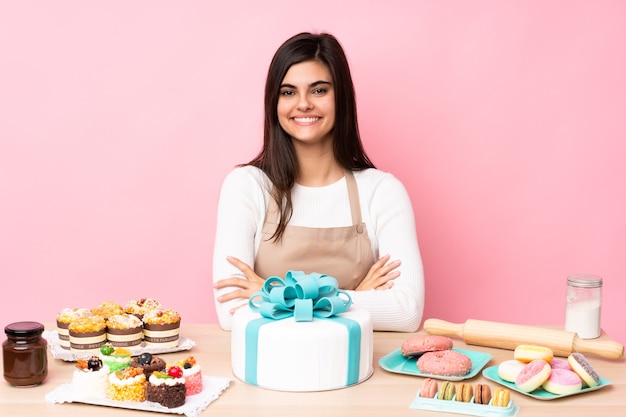 Chef de confeitaria com um grande bolo em uma mesa sobre uma parede rosa isolada com os braços cruzados e olhando para a frente