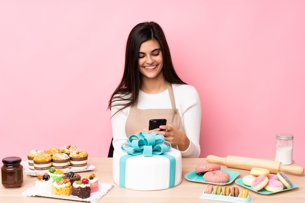 Chef de confeitaria com um grande bolo em uma mesa sobre uma parede rosa isolada enviando uma mensagem com o celular