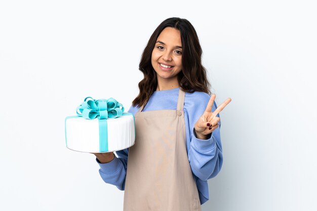 Chef de confeitaria segurando um grande bolo sobre uma parede branca isolada, sorrindo e mostrando o sinal da vitória