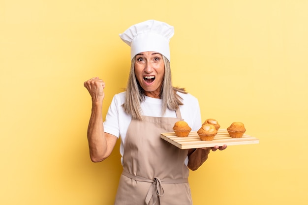 Chef de meia-idade gritando agressivamente com uma expressão de raiva ou com os punhos cerrados comemorando o sucesso e segurando uma bandeja de muffins