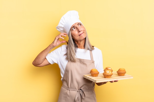 Chef de meia-idade se sentindo perplexa e confusa, coçando a cabeça e olhando para o lado segurando uma bandeja de muffins