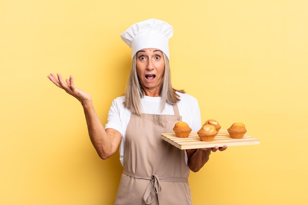 Chef de meia-idade sentindo-se extremamente chocada e surpresa, ansiosa e em pânico, com uma aparência estressada e horrorizada e segurando uma bandeja de muffins