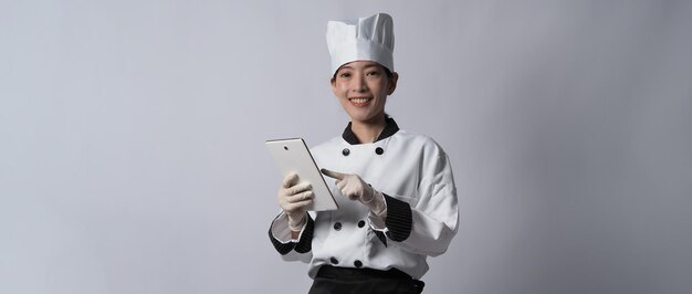 Chef de mulher asiática segurando um smartphone ou tablet digital e recebeu o pedido da loja online ou do aplicativo do comerciante. ela sorrindo em uniforme de chef e em pé no fundo branco. Comerciante de comida online.