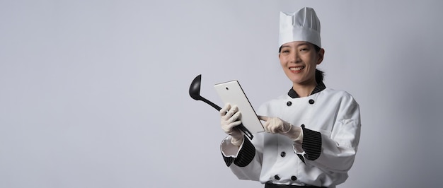 Chef de mulher asiática segurando um smartphone ou tablet digital e recebeu o pedido da loja online ou do aplicativo do comerciante. ela sorrindo em uniforme de chef e em pé no fundo branco. comerciante de comida online.