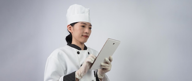Chef de mulher asiática segurando um smartphone ou tablet digital e recebeu o pedido da loja online ou do aplicativo do comerciante. ela sorrindo em uniforme de chef e em pé no fundo branco. Comerciante de comida online.
