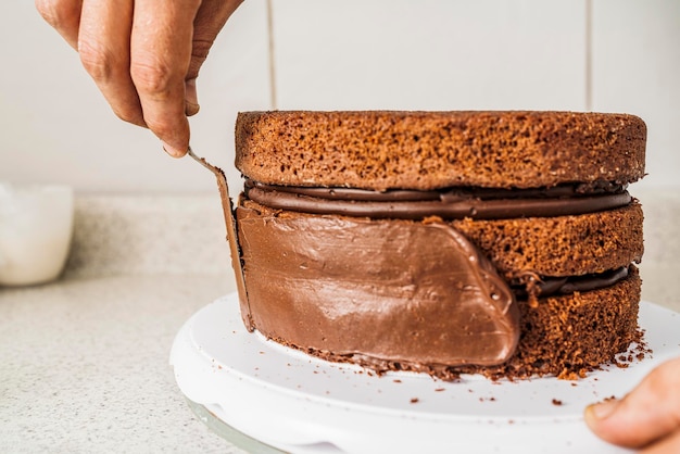 Chef de pastelaria espalhando creme de chocolate nas bordas de um bolo de chocolate dentro de casa