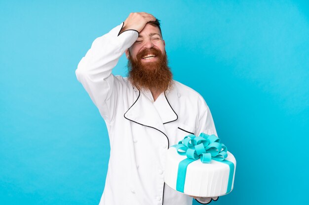 Chef de pastelaria ruiva com barba longa, segurando um bolo grande sobre parede azul isolada, percebeu algo e pretendia a solução